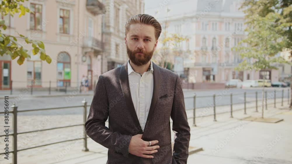 Attractive bearded businessman standing look at camera serious in old city at sunlight. Elegant handsome confident man. Entrepreneur leader successful. Slow motion Attractive bearded businessman standing look at camera serious in old city at sunlight. Elegant handsome confident man. Entrepreneur leader successful. Slow motion