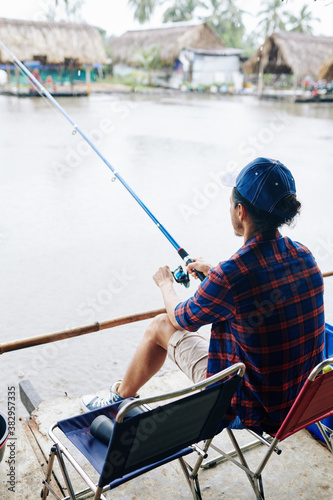 Mature man sitting on folding chair and fishing in lake with new spinning, view from the back