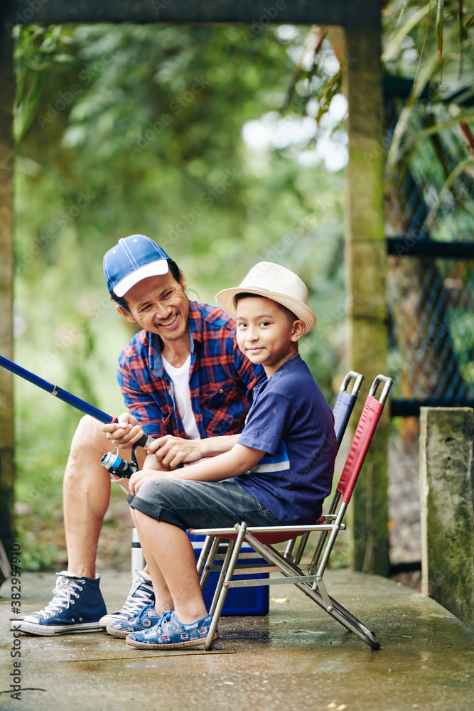 Cute smiling little boy sitting on folding chair and fishing with spinning with his dad Cute smiling little boy sitting on folding chair and fishing with spinning with his dad