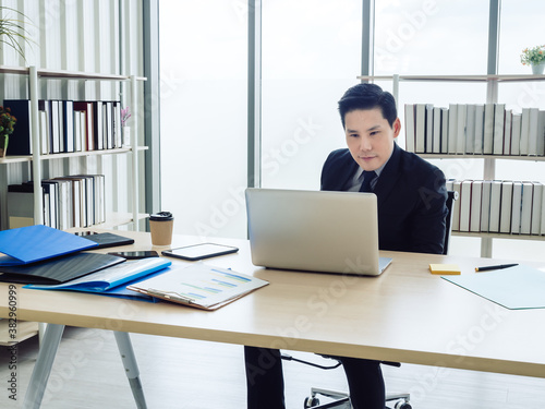 Asian businessman in suit sitting at table and working on laptop computer in office. Manager watching and typing on laptop computer in well-lit workplace. Business, technology and finance concept.