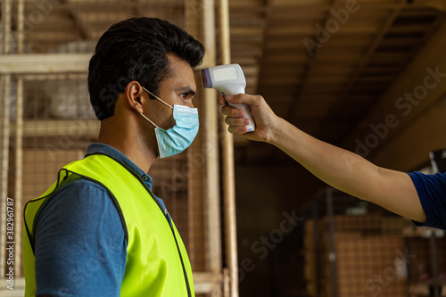 Young Indian factory warehouse worker wearing face mask getting body temperature check with infrared thermometer scanner before working. Security measure at workplace in Coronavirus Covid 19 pandemic