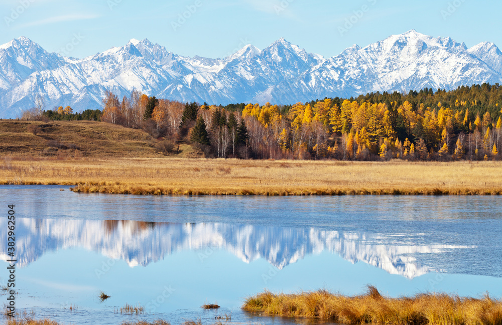 Yellowed forest and snow-capped mountain peaks are reflected in a calm river covered with a crust of ice on a sunny autumn day. Beautiful landscape, natural background, autumn travel