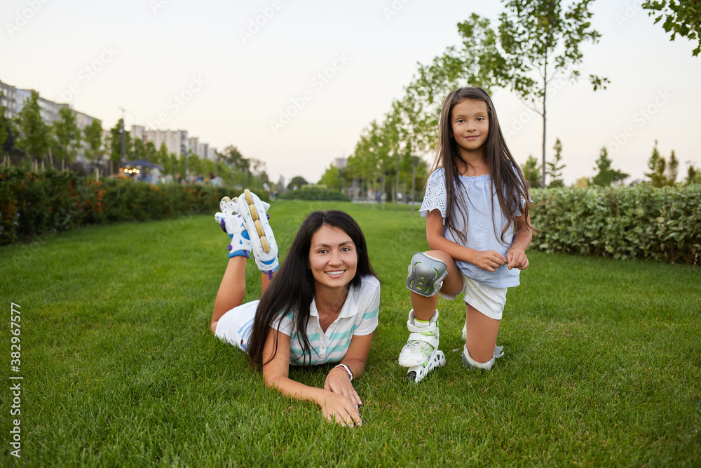 Fototapeta premium mother and daughter in roller skates are sitting on grass and having fun in park.