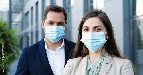 Portrait of Caucasian young man and woman in business style and in medical masks standing at street and looking at camera. Businessman and businesswoman outdoors during coronavirus pandemic.