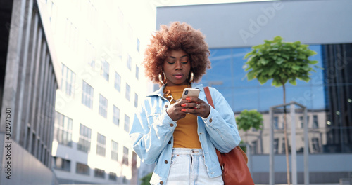 African American young stylish curly woman tapping or scrolling on smartphone and standing at city street. Beautiful female texting message on mobile phone and chatting. Outside. photo