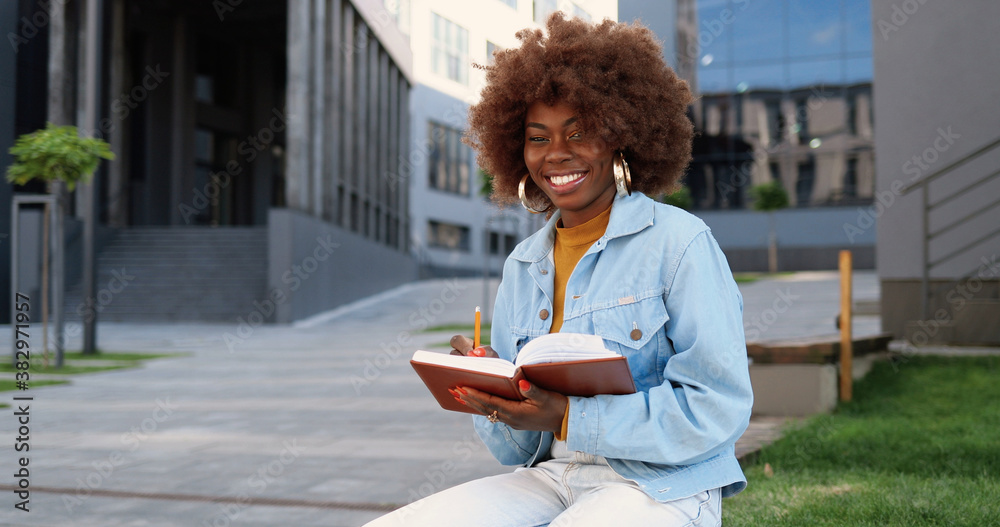 Portrait of young curly and pretty African American woman sitting on bench, reading textbook, making marks and smiling to camera. Portrait of young curly and pretty African American woman sitting on bench, reading textbook, making marks and smiling to camera.