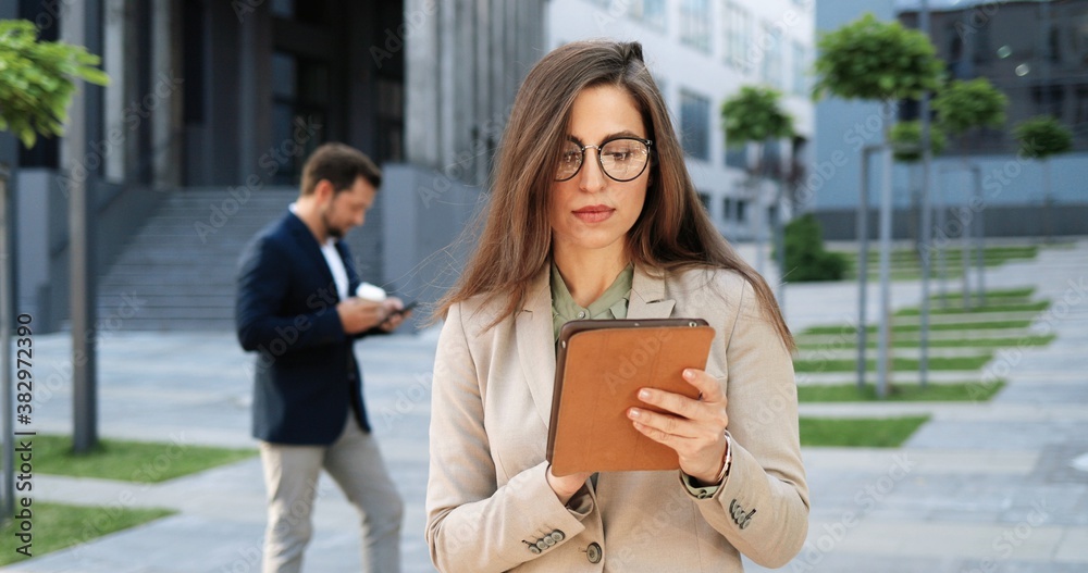 Fototapeta premium Caucasian attractive young stylish woman tapping or scrolling on tablet device in town at street. Beautiful female texting message and using gadget computer. Businesswoman in glasses working outdoor.