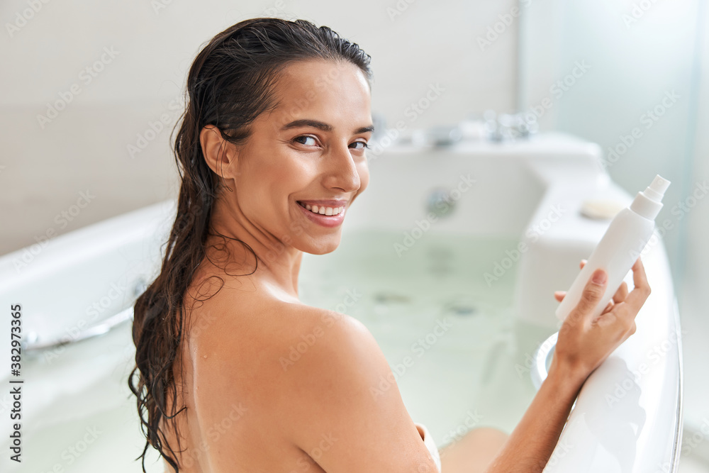 Obraz premium Cheerful young woman holding spray bottle of lotion while taking bath