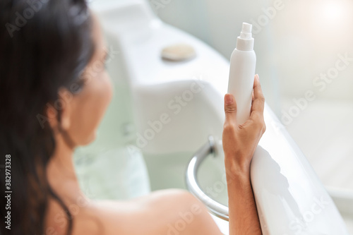 Young woman holding spray bottle of lotion while taking bath