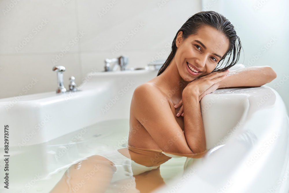 Fototapeta premium Cheerful young woman lying in white bathtub at home