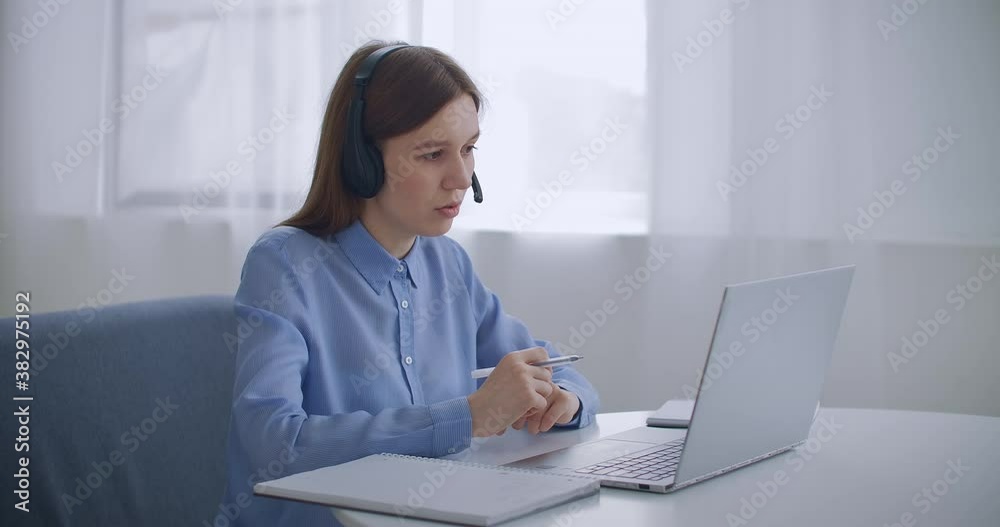 female specialist of call-center is talking by headset and looking at display of laptop, portrait indoors, working remotely female specialist of call-center is talking by headset and looking at display of laptop, portrait indoors, working remotely