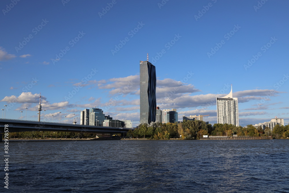 Der DC Tower vor der UNO City auf der sogenannten Donauplatte ist vor der Reichsbrücke und der Wiener Donau zu sehen. Es ist leicht bewölkt bei schönem blauen Himmel.
