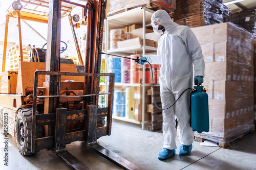 Worker in sterile uniform with rubber gloves holding sprayer with disinfectant and spraying around warehouse. Corona outbreak concept. © dusanpetkovic1