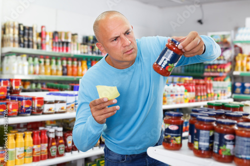 Positive male purchaser with shopping list choosing food products in grocery