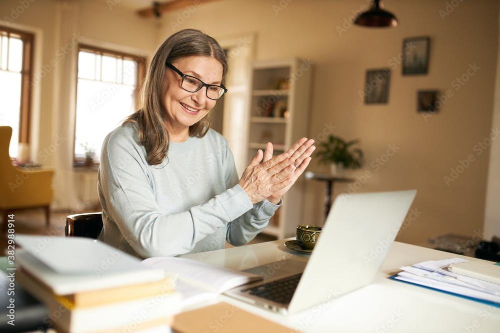 Technology, online communication and electronic gadgets concept. Positive joyful female pensioner in spectacles using laptop talking to family via webcam video conference chat, smiling, clapping hands Technology, online communication and electronic gadgets concept. Positive joyful female pensioner in spectacles using laptop talking to family via webcam video conference chat, smiling, clapping hands
