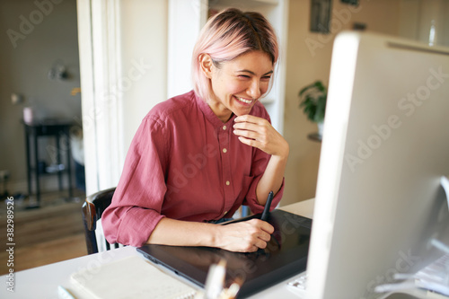 Indoor shot of cheerful happy young female with stylish pinkish hair laughing while working from home, sitting at desk with computer and graphic tablet, retouching images or drawing animation