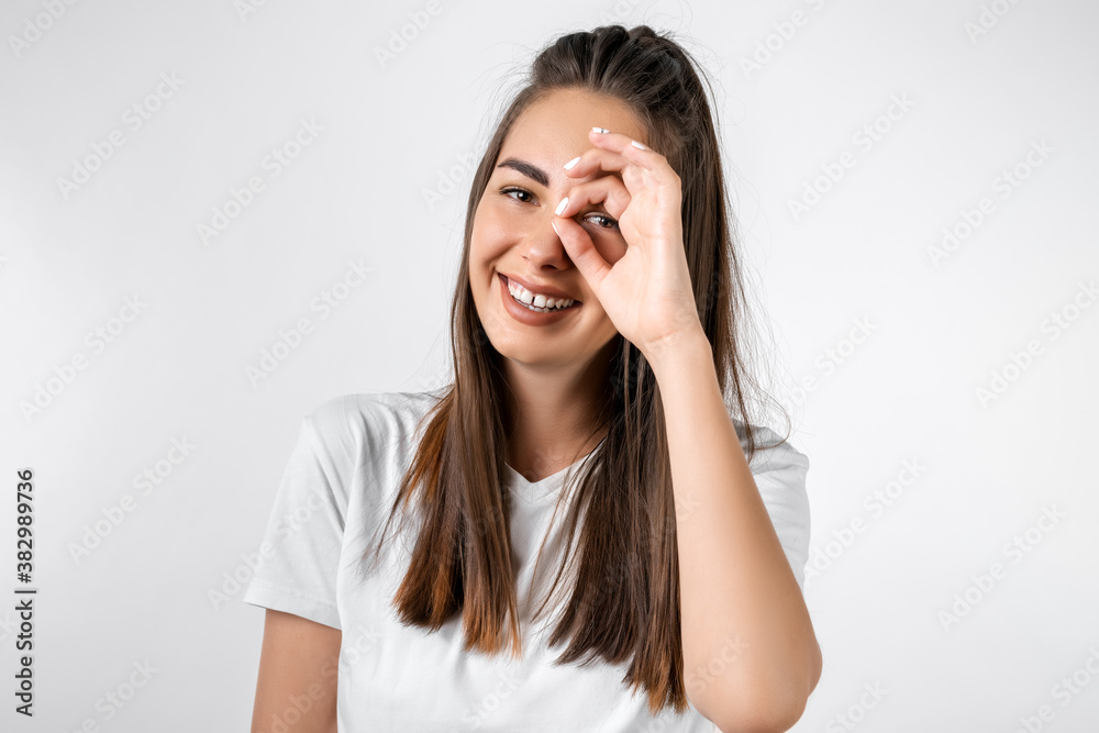 Cheerful smiling girl looking at the camera through fingers in OK gesture. Studio shot white background Cheerful smiling girl looking at the camera through fingers in OK gesture. Studio shot white background