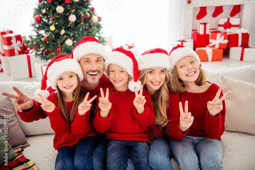 Full family x-mas vacation. People mommy mom dad daddy three little preteen kids sit couch make v-sign wear santa claus cap in house indoors with noel eve tinsels