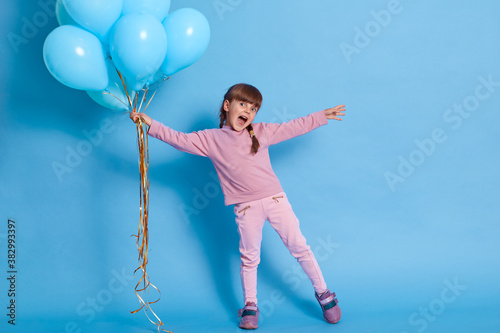 Girl with bunch of helium balloons spreading hands aside and yelling something happily, wearing casual rosy jumper, trousers and sneakers against blue background, cute child at party.