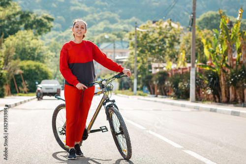 Sports and activity. A pretty woman in red sportswear is pushing a Bicycle along an empty road. Copy space