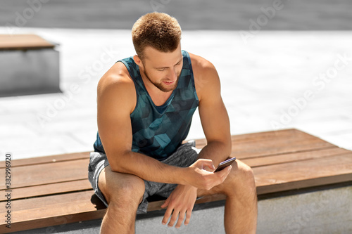 fitness, sport and technology concept - happy smiling young man with smartphone sitting on bench outdoors
