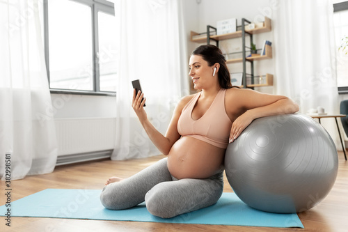 pregnancy, sport and fitness concept - happy smiling pregnant woman with smartphone, earphones and fitball exercising at home