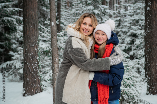 Beautiful blonde mom in a fur coat and her teenage daughter pose for a photo among the trees in winter.