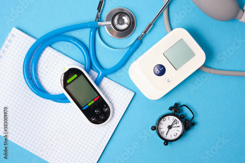 Digital blood pressure monitor, glucometer, stethoscope, notepad and alarm clock on blue background top view