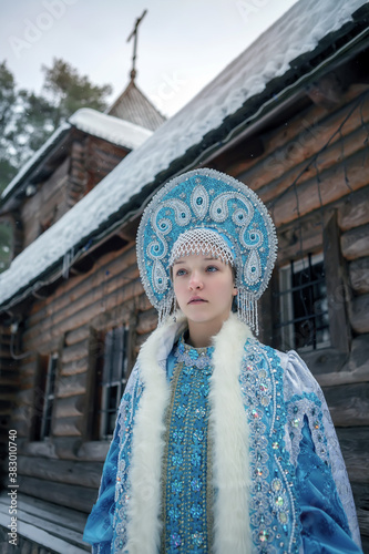 A girl in a Snow Maiden costume stands against the background of a winter forest and a wooden house. Winter, December. Russian Christmas character Snegurochka. New Year.