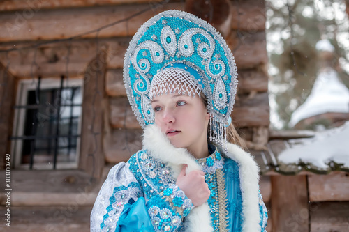 A girl in a Snow Maiden costume stands against the background of a winter forest and a wooden house. Winter, December. Russian Christmas character Snegurochka. New Year.