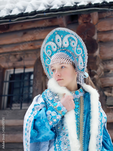 A girl in a Snow Maiden costume stands against the background of a winter forest and a wooden house. Winter, December. Russian Christmas character Snegurochka. New Year.