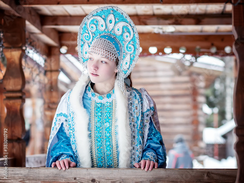 A girl in a Snow Maiden costume stands against the background of a winter forest and a wooden house. Winter, December. Russian Christmas character Snegurochka. New Year.