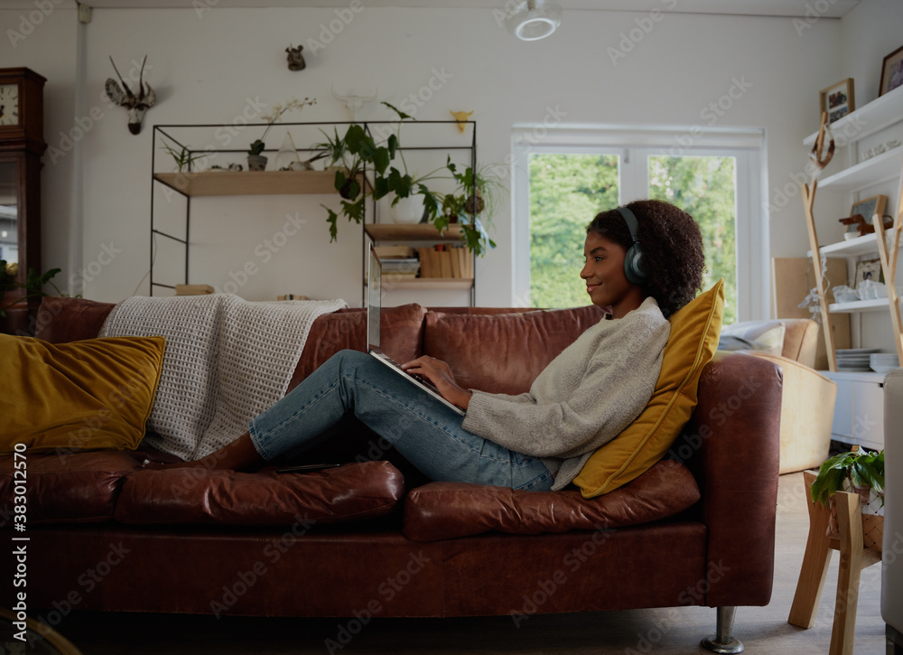 Side view of young woman relaxing at home watching webinar and typing with headphones sitting on couch Side view of young woman relaxing at home watching webinar and typing with headphones sitting on couch