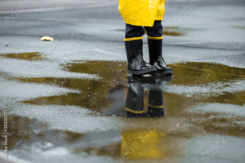 Boy in yellow waterproof cloak and rubber boots standing in a puddle outdoors in the rain in autumn.