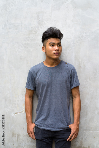 Young Asian man with curly hair thinking against concrete wall outdoors