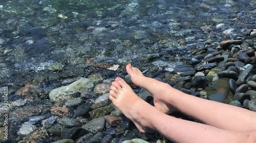 Legs and feet of a child sitting on a pebble sea shore and relaxing, salt water rolls over his legs