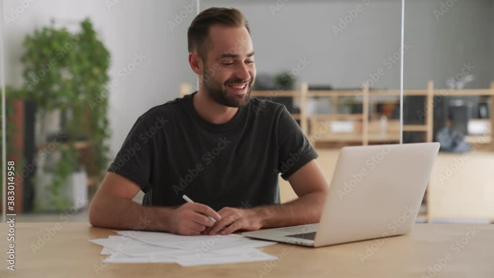 Young positive happy man indoors in office using laptop computer and writing notes