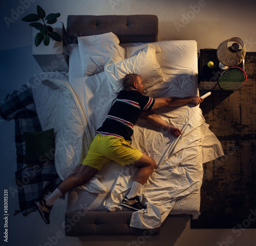 Dreamlike. Top view of young professional tennis player sleeping at his bedroom in sportwear with racket. Loving his sport even more than comfort, watching match even if resting. Action, motion, humor