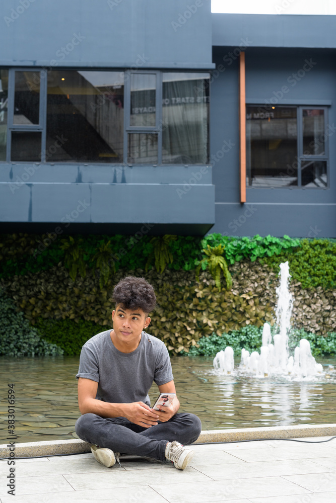 © Ranta Images - Young Asian man with phone thinking and sitting outside modern building © Ranta Images - Young Asian man with phone thinking and sitting outside modern building