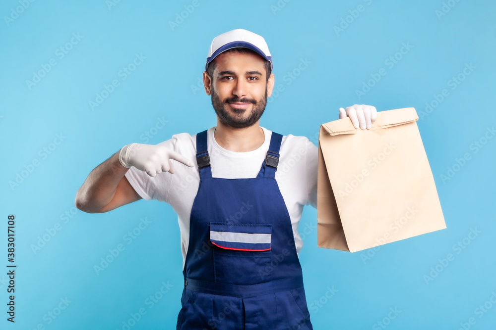 Look at bag! Portrait of happy smiling handyman in overalls and gloves pointing at ordered parcel. Courier delivering food in paper package, post mail services. studio shot isolated on blue background