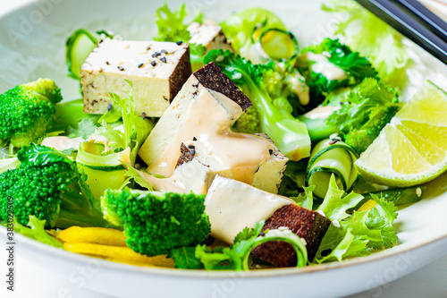 Green Asian salad with broccoli, smoked tofu and peanut dressing in white bowl, white background.