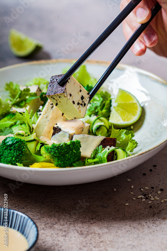Green vegan salad with broccoli, smoked tofu and tahini dressing in white bowl, dark background.