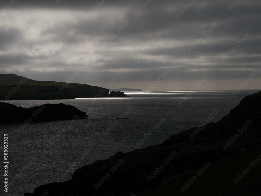 In black and white, a moody, threatening view of sunlight on the water across Magnus Bay from hillside at Islesburgh near Mavis Grind,  Shetland, UK.