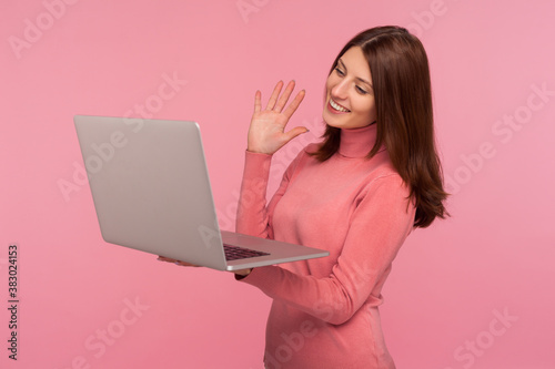 Smiling brunette woman beauty blogger saying hello to her subscribers recording video for blog on laptop, video call. Indoor studio shot isolated on pink background