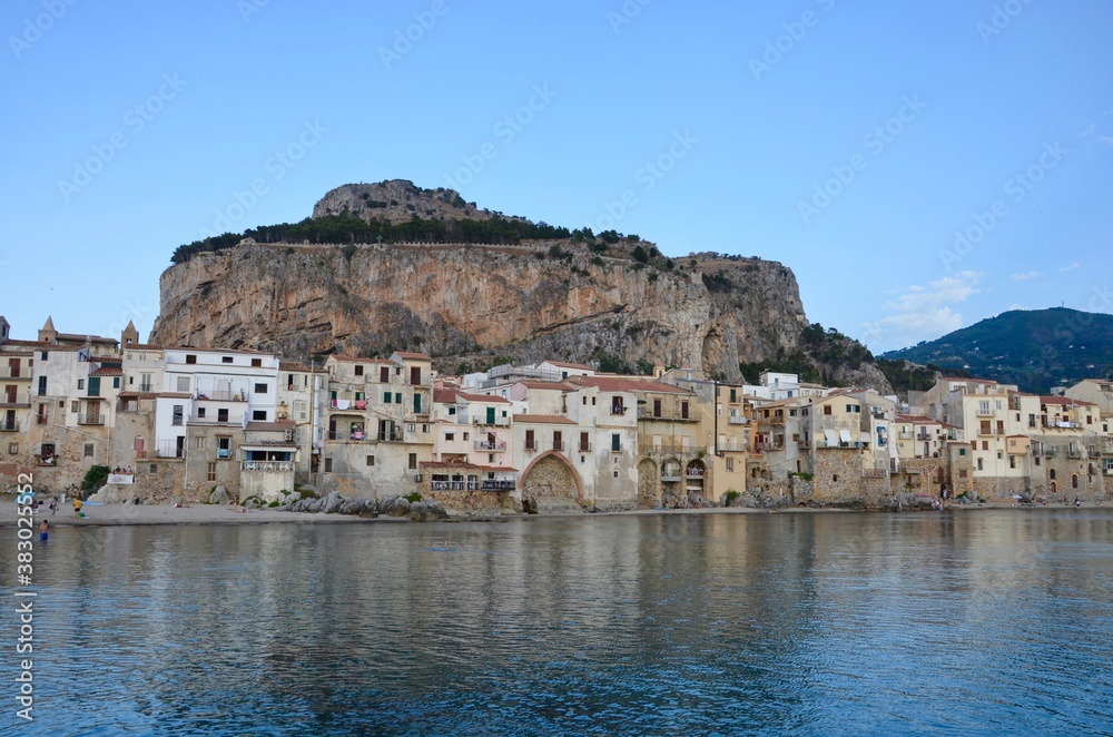 Coastal town Cefalu in Sicily, view from quay to the city beach and castle rock, medieval buildings in the background, late afternoon in summer, a sunny day