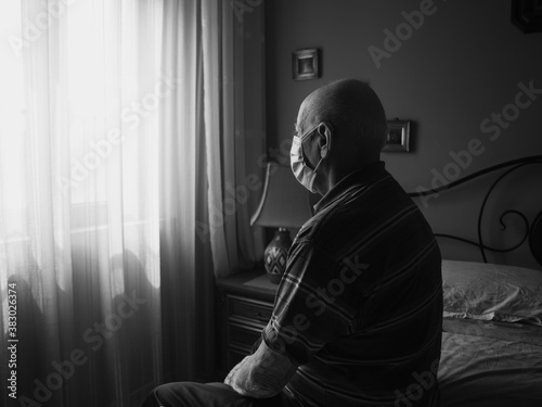 COVID-19 Black and white profile view of an old man wearing an antivirus mask sitting on the bed looking at the window  © MarcoDiStefano