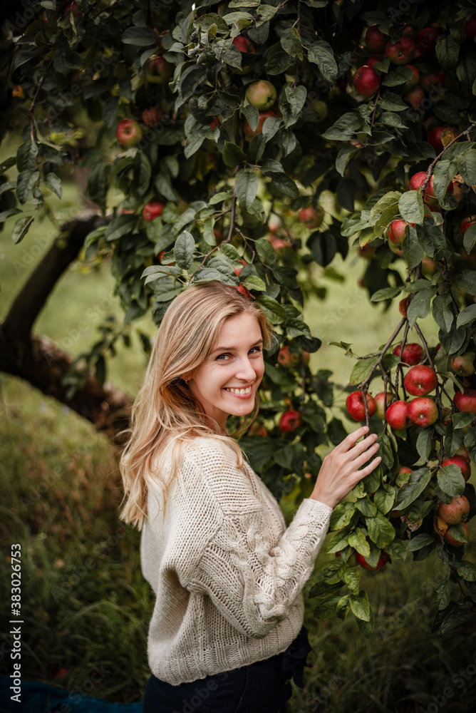 Woman smiling to camera while posing next to apple tree wth red apples on a warm autumn evening