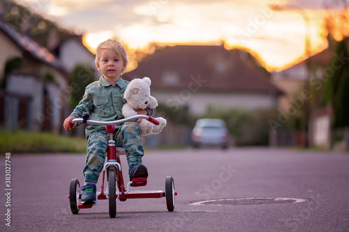 Toddler child, blond boy, riding tricycle in a village small road on sunset