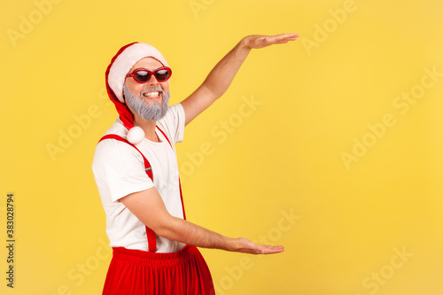 Happy grey bearded man in santa claus hat and pants with suspenders showing free space for your advertising, looking at camera with toothy smile. Indoor studio shot isolated on yellow background