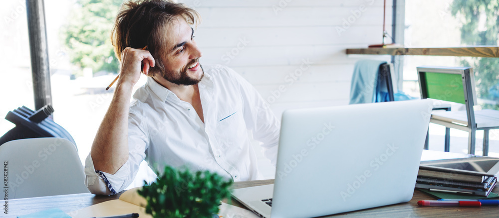 Cheerful thoughtful man with stubble sitting at home and working remote using laptop. Private house workplace. Wide screen, panoramic Cheerful thoughtful man with stubble sitting at home and working remote using laptop. Private house workplace. Wide screen, panoramic